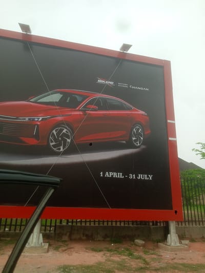 The image shows a large outdoor advertisement in Kubwa, Nigeria. The advertisement is for Changan Motors, specifically featuring a red car. The advertisement includes the text "1 APRIL - 31 JULY". The billboard has a red frame. There are two lights on the top of the billboard. The billboard is situated next to a fence and vegetation. The sky is overcast. A car doorframe is visible on the left side of the image, partially obscuring the view.