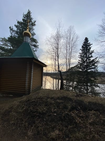 The image depicts a wooden chapel with a green roof and a golden dome topped with a cross. The chapel is surrounded by trees and is partially obscured by a dirt bank covered in dry vegetation. The sky is overcast. The exact location within the city of Pepelyshi, Russia is not discernable from the image.