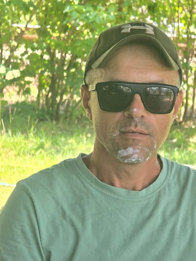 A close-up, head-and-shoulders portrait of a man wearing a green t-shirt, a green baseball cap with an Under Armour logo, and black sunglasses. He has facial hair and white foam around his mouth and on his chin. The background is blurred and shows greenery, likely trees and grass, suggestive of an outdoor, possibly park-like setting in Kresna, Bulgaria. The lighting appears to be natural daylight.
