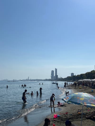 The image depicts a busy beach scene on a clear, sunny day, likely in Germasogeia Municipality, Cyprus, given the presence of modern high-rise buildings in the background that are characteristic of the area. The sea is calm with gentle waves lapping at the sandy shore. Numerous people are enjoying the water and the beach. In the water, individuals of various ages are swimming, wading, and playing. On the beach, there are groups of people relaxing under umbrellas, with some children playing with sand toys. A colorful beach umbrella is prominently featured in the foreground. In the distance, a hazy skyline is visible with several tall buildings and a harbor or port area, suggesting an urban coastline. The overall atmosphere is one of leisure and recreation.