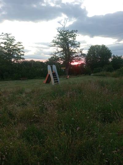 The image depicts an outdoor scene at dusk in Ароматновское сельское поселение, Ukraine. A playground with a slide and a seesaw is visible in a grassy area overgrown with tall weeds and wild grasses. Several trees, some bare and others with foliage, frame the scene, with a dense line of trees in the background. The sky is cloudy, with the setting sun casting a reddish glow. Power lines are visible in the distance. The overall impression is one of a slightly neglected, rural park or playground.