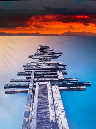 The image depicts a wooden pier stretching into a calm body of water. The pier is constructed from weathered, rough-hewn planks, arranged in a staggered, almost abstract fashion, with some sections appearing to jut out precariously. At the base of the pier, a set of wooden steps leads upwards. The water is a deep, serene blue, reflecting the clear sky above. In the distance, a range of mountains is silhouetted against a dramatic sunset. The sky is ablaze with fiery oranges and reds, transitioning to darker, somber tones towards the top of the frame. There are no people visible in the image. The scene is serene and evocative, with the starkness of the wooden structure contrasting with the vibrant, natural beauty of the sunset and the landscape. The location in the city of Salsk, Russia, is not discernible from the visual elements presented in the image.