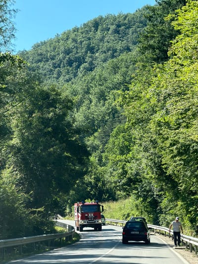 In Troyan, Bulgaria, on a sunny day, a red fire truck with "IVECO" and "MAGIRUS" markings is driving on a paved road that curves to the left, bordered by a metal guardrail and dense green foliage on both sides. The road is also occupied by a black SUV traveling ahead of the fire truck and a man in a white hat, white t-shirt, and dark pants walking along the shoulder of the road to the right. The background consists of a steep, tree-covered mountain under a clear blue sky.