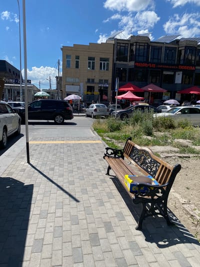 This image depicts a street scene in Gyumri, Armenia, on a sunny day, indicated by the clear blue sky and strong shadows. The perspective is from a sidewalk looking down the street, with the buildings on the left and a pathway on the right.

The main subjects include several cars parked along the street, some with people visible inside or nearby. A dark blue car is prominently featured. On the sidewalk, there is a decorative bench with a wooden seat and ornate black metalwork. A yellow and blue packaged item rests on the bench, partially obscured. The pathway is paved with grey bricks, with a yellow tactile paving strip running along its edge. Greenery and grass grow in patches between the pavement and the buildings.

The buildings on the left appear to be commercial establishments. One building has a sign with Cyrillic script that reads "Must-SUPERMARKET ПОБЕДА" and another with "U-Z 3 LH". Another building has signs like "Baby Beer" and "ԱՐՏ ՇՈՓ". Red and pink umbrellas or awnings are visible in front of some of the businesses.

The overall scene suggests a typical day in a town or city, with people going about their business. The weather is fair, and the time of day appears to be mid-morning or afternoon, given the angle of the shadows. The presence of parked cars, shops, and a public bench suggests an urban environment.