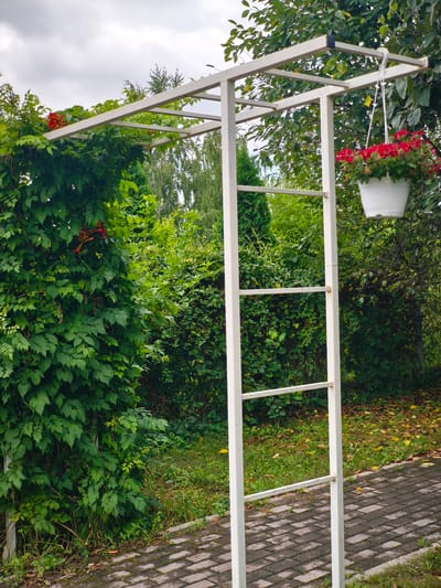 The image depicts an outdoor scene in a garden or park setting, likely in Nevinnomyssk, Russia, based on the provided context. The central focus is a white metal pergola structure, which serves as an archway. Lush green foliage, predominantly from a climbing plant, heavily drapes over the left side and top of the pergola, with some orange trumpet-shaped flowers visible among the leaves.
To the right of the pergola, a white hanging basket filled with vibrant red flowers is suspended from the structure. The ground in front of the pergola is paved with dark grey and reddish-brown rectangular bricks arranged in a pattern, creating a pathway. Beyond the paved area and the pergola, a dense green hedge forms a backdrop, with trees and other vegetation visible further in the distance. The sky above is overcast with grey clouds. The overall scene suggests a well-maintained outdoor space, possibly a garden or a park, with a focus on greenery and floral decorations. There are no people or animals visible in the image.