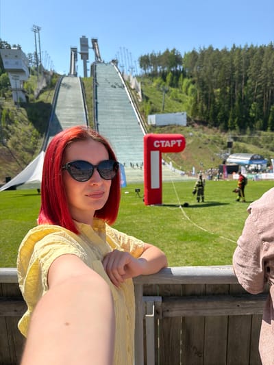 A woman with red hair and sunglasses is posing in front of a ski jumping venue. The venue includes a large ski jump ramp and a "СТАРТ" (START) sign. The location is in Прикамский, Russia, as suggested by the venue's presence. The woman is wearing a yellow shirt and holding onto a wooden fence. Other people and structures are visible in the background.