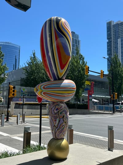The image depicts a brightly colored, abstract sculpture composed of stacked, rounded shapes. The sculpture is positioned outdoors on a sunny day, with a clear blue sky overhead. In the background, modern city buildings and trees are visible, suggesting an urban environment.

The sculpture's lowest element is a large, gold, spherical base. Above this sits a vase-like shape with a purple and swirling multicolor pattern. The next element is a horizontally oriented, flattened oval covered in a multitude of thin, colorful stripes. The topmost element is an elongated, egg-like shape with bold, wavy stripes in red, yellow, blue, purple, black, and white.

The surrounding area features a paved sidewalk with a brick pattern, low wooden planters with grass, and metal bollards. Across a street, a building with a large "BU" logo is visible, along with traffic lights and street signs. One sign indicates "STATION SQUARE". Another sign shows "METRO at metro". The presence of "STATION SQUARE" and the architectural style of the surrounding buildings are indicative of the area near the Metrotown transit station and mall complex in Burnaby, British Columbia, Canada. The scene is devoid of people.