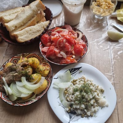 A close-up overhead shot shows a meal spread on a light brown wooden table, possibly in an apartment kitchen in Aktobe, Kazakhstan. In the upper left, a dark bowl contains sliced white bread. Adjacent to it, a clear plastic cup holds a white creamy substance. To the right, a glass bowl filled with a crispy yellow food item sits near a partially squeezed yellow pouch and a knife. A segment of white onion is visible on the table near the knife.
In the mid-left, a round, patterned bowl holds a salad of chopped tomatoes and white onions, mixed with a creamy dressing. Below that, another patterned bowl presents roasted whole potatoes and pieces of meat, garnished with fresh dill and sliced white onions. In the lower right, a white plate contains white rice mixed with green peas and dill, also with slices of white onion. The overall scene depicts a home-cooked meal with various dishes, suggesting a casual dining setting.