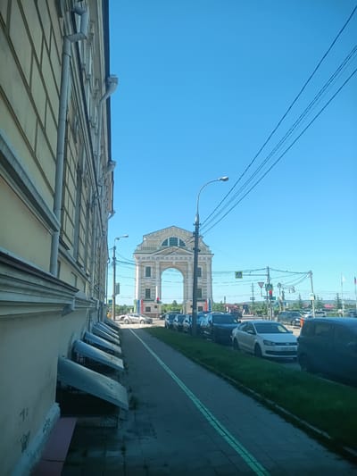 The image depicts a street scene in Irkutsk, Russia, on a clear, sunny day. The time of day appears to be late morning or early afternoon, given the bright sunlight and distinct shadows. The foreground is dominated by the side of a light-colored, classical-style building with a row of grey, angled awnings along its base. The building's architecture suggests it's an older structure, possibly a historical landmark or a well-maintained government building.
The street is paved with cobblestones, and a sidewalk runs alongside the building, marked with double green lines. Several cars are parked along the street, and more are in motion. Among the vehicles visible are a white Volkswagen Polo with the license plate E031XX 38, and a dark-colored SUV.
In the background, a prominent archway structure stands out against the bright blue sky. This archway, also light-colored and grand in design, features decorative elements and a large arched opening. It appears to be a significant landmark within the city. Traffic lights and street lamps line the street, indicating an urban environment. The presence of pedestrian crossing signs further suggests this is a busy thoroughfare. The overall atmosphere is calm and ordinary, capturing a typical day in this Russian city. No specific activity or event is clearly identifiable beyond normal urban traffic and pedestrian flow.