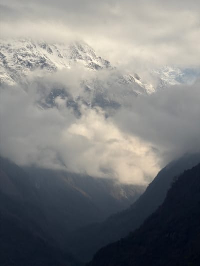 The scene depicts a deep mountainous valley and peaks, consistent with a view from the vicinity of Ghandruk, Nepal. The upper background features large, snow-capped mountain peaks, partially obscured by dense, turbulent grey and white clouds. Below these high peaks, a deep valley or gorge is visible, with steep, dark, shadowed slopes defining the lower and mid-sections of the image. A distinct shaft of sunlight penetrates the cloud cover in the middle of the frame, illuminating a section of the clouds or a distant valley wall. No people, animals, structures, or any discernible man-made objects are present within the frame. The environment is rugged, natural, and characterized by dramatic atmospheric conditions and mountainous terrain.