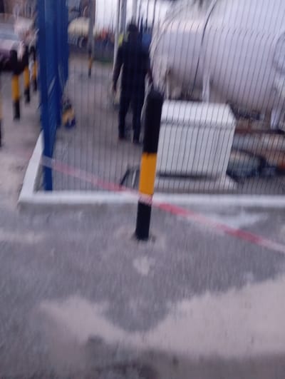 The image depicts an outdoor industrial area, likely within a facility in Port Harcourt, Nigeria, given the context. In the foreground, a grey, textured ground surface is visible, interspersed with patches of lighter colored material, possibly sand or dust. A black and yellow striped bollard stands prominently in the mid-ground, partially obscuring a red and white caution tape that stretches diagonally across the frame. Beyond the bollard, a chain-link fence separates the foreground from the background. Through the fence, a large, white cylindrical tank is visible, suggesting the presence of an industrial or storage facility. To the left of the tank, a white, rectangular structure with a ribbed surface is present, possibly a control box or storage unit. In the far background, a person, appearing to be male and wearing dark clothing, is walking away from the camera. Other figures and objects are blurred and indistinct due to the image's low resolution and motion blur. The overall impression is that of an active industrial site.
