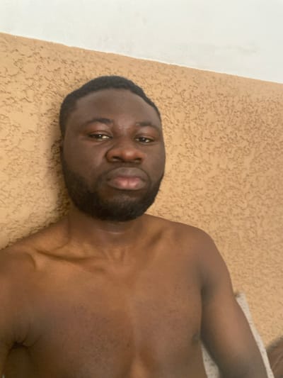 This image depicts an adult male, identified as being in Udu, Nigeria. The subject, of dark complexion, has short dark hair and a dark, trimmed beard and mustache. He is shirtless, with his bare chest and shoulders visible, facing directly towards the viewer with a neutral expression and slightly pursed lips. His gaze is directed forward. The background consists of a light brown or tan wall featuring a stucco-like texture, which meets a plain white ceiling in the upper portion of the frame. The scene is illuminated by uniform lighting, indicating an indoor environment.