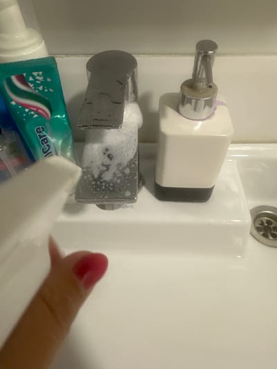 In an indoor scene, a white sink is visible with a chrome faucet that has foam around its base. To the right of the faucet is a white soap dispenser with a chrome pump. A tube of toothpaste with green, pink, and white branding is placed to the left of the faucet. A person's hand with pink fingernail polish is holding a white spray bottle, with the nozzle pointed towards the faucet and sink, suggesting an action of spraying. The background appears to be a tiled wall. This scene is located in Novoaltaysk, Russia.