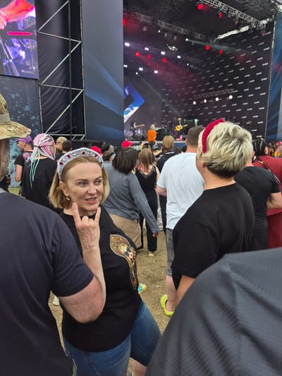 The image depicts an outdoor music festival with a stage in the background. A woman in the foreground is wearing a black t-shirt with a graphic print on it and a silver headband with red embellishments. She is making a "rock on" hand gesture with her right hand. She appears to be speaking or singing, with her mouth open. To her left, a person wearing a dark blue t-shirt is partially visible. Behind the woman, a crowd of people are gathered, facing the stage. Some are wearing colorful headwear, including pink braids and a red headband. The stage has a large screen and lighting. The ground is covered with dry grass. The overall atmosphere suggests a lively event. The provided location information, "city of Lagunovo, Russia", is stated as fact for this description.