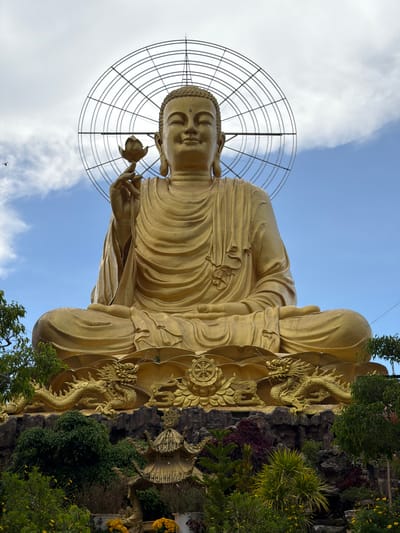 This image captures a monumental golden statue of a seated Buddha against a bright blue sky with scattered clouds. The Buddha holds a lotus flower in its right hand, and a circular halo with radiating lines is positioned behind its head. The statue is adorned with intricate drapery. It rests on a multi-tiered platform featuring golden dragon sculptures, decorative patterns, and rocky elements. At the base of the statue, there is a lush, green landscape with various plants and yellow flowers. A small, multi-tiered golden pagoda-like structure is also visible in the foreground, partially obscured by foliage. The overall scene suggests a significant religious or cultural site, likely a temple or monument located in Lâm Viên - Đà Lạt, Vietnam. The lighting indicates it is daytime, likely a clear or partly cloudy day.