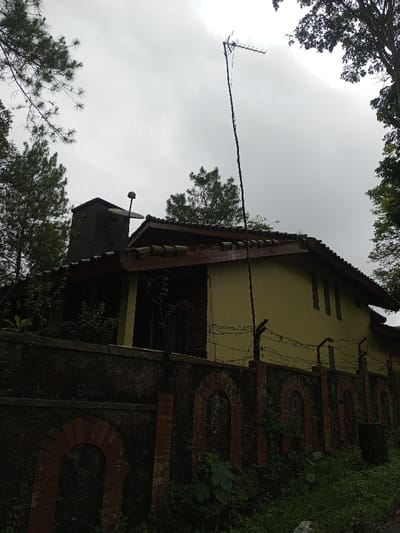 The image displays an exterior view of a two-story building and its perimeter wall, located in Ciherang, Indonesia, observed from a low angle. The building features pale yellow main walls and a brown tiled roof with dark brown eaves. On the left side of the roof, a dark, block-like chimney is visible, accompanied by a small, dark, spherical object mounted nearby. Three narrow, dark vertical window openings are present on the upper right facade of the building. A tall, slender dark pole, topped with a directional television antenna, stands to the right of the structure, extending significantly above the roofline.

The foreground is dominated by a dark, textured concrete or stone perimeter wall, partially covered by green vegetation. This wall incorporates multiple brick pillars with arched recesses. Strands of barbed wire are secured horizontally along the top of several dark metal posts embedded in the wall, serving as a security measure. Lush green foliage, including tall pine-like trees on the left and broadleaf trees on the right, frames the upper portions of the scene. Overgrown grass, weeds, and various leafy plants populate the ground directly in front of the wall. The sky is overcast and light grey. No individuals or specific actions are discernible within the frame.