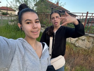 Two young women pose for a selfie outdoors. One is wearing a grey zip-up hoodie and has her hair in a bun; she has an earpiece in her left ear. The other is wearing a black zip-up jacket with a white crossbody bag and is sticking out her tongue while making a hand gesture. They are standing in front of overgrown vegetation and a dilapidated structure with a rusty fence. The surroundings suggest a rural or semi-rural environment. Based on the prompt, the location is Karavelovo, Bulgaria.