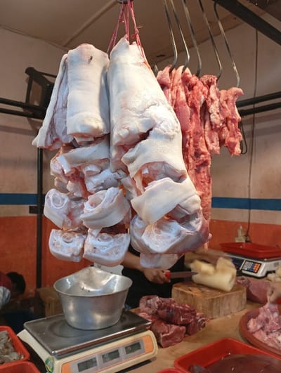 The image shows a butcher shop or market stall in Kutawaringin, Indonesia, at the location of latitude: -6.953, longitude: 107.524. The main subject is a variety of raw meat cuts, including pork legs and other cuts, hanging from a metal rack, ready for sale. The meat is pale pink and white, and the cuts are arranged in a neat, organized manner. The meat is displayed on a metal frame and is partially obscured by a white metal container. Other cuts of meat can be seen on the table below, including red meat and what appears to be pork belly. There is a scale in the bottom left of the image, suggesting that the meat is being weighed for sale. A person's hand can be seen in the bottom center of the image, partially obscured by the meat. There is also a white rectangular object that appears to be a plastic cutting board. The lighting is bright and the image is taken from an angle that shows the entire stall.