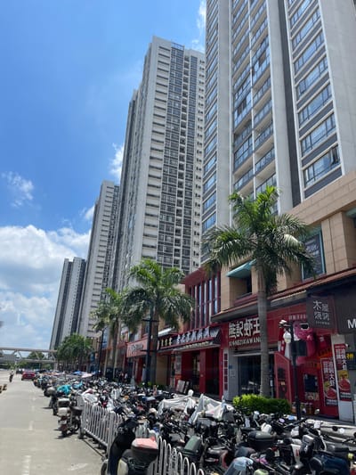 This is a street-level, eye-level, outdoor photograph taken on a sunny day in Panyu District, China. The image captures a row of tall, modern apartment buildings, stretching upwards against a clear blue sky with scattered white clouds. In the foreground, a street is lined with numerous parked motorcycles, most of them covered with white protective sheets. A white metal fence separates the sidewalk from the motorcycle parking area. Along the street, a series of commercial establishments are visible, featuring brightly colored signage, predominantly in red and white, with Chinese characters. Palm trees are interspersed along the sidewalk, providing some shade. In the background, a traffic bridge with a rail system is visible. A few cars are seen on the road, and a lone person is on a motorcycle in the distance. The architecture of the buildings is characterized by repetitive window patterns and balconies. The overall scene depicts a busy urban environment with a strong focus on residential high-rises and street-level commerce.