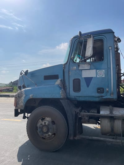 A light blue, weathered heavy-duty truck with significant paint wear and exposed metal on the front fenders is depicted on a paved road under a bright, clear blue sky with scattered clouds. The truck's cab is visible from the front-left side, revealing an open driver's door, with a person partially discernible within the driver's seat. Visible details on the cab include an American flag decal and two other white stickers on the driver's door, one featuring a circular logo with green and red elements. A prominent blue inverted triangular marking is also present on the door. The front-left wheel and tire are clearly shown, displaying accumulated dirt and wear. In the mid-ground, power lines stretch horizontally across the frame. The background to the left features blurred vegetation and a dark, possibly unfinished or dilapidated structure. The scene is located in the city of Ibeju, Nigeria.