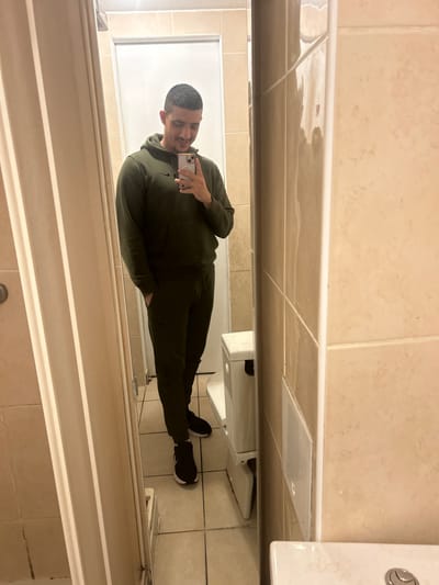 A man stands in a bathroom, taking a mirror selfie with his smartphone. He is wearing a dark green tracksuit and black sneakers. The bathroom features beige tiled walls and flooring. A toilet is visible on the right side of the image. The overall scene is typical of an everyday moment captured in a casual photograph.