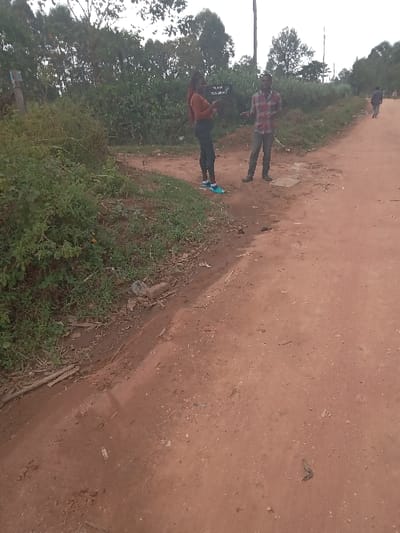 The image shows two people standing on a dirt road, located in Burundu, Kenya, at the coordinates (latitude: 0.491, longitude: 34.79). The woman on the left is wearing a long-sleeved orange shirt and dark blue jeans. She is looking at a sign that reads "Village Settlement" in black lettering. The man on the right is wearing a red and black plaid shirt and dark blue pants. He is looking at the woman. The background shows a dirt road, green vegetation, and trees. The scene appears to be a casual interaction between two individuals in a rural area.