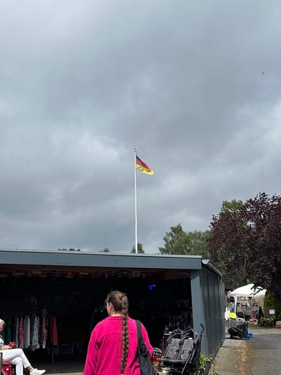 The image depicts an outdoor scene under a heavily overcast, grey sky in Deinste, Germany. In the upper middle-right, a German flag, displaying horizontal black, red, and gold stripes, flies from a tall white flagpole. Below the flag, a dense cluster of green trees and a tree with dark reddish-purple foliage are visible.

The foreground is dominated by a dark grey, corrugated metal structure, appearing to be a market stall or shop, with its front open. The opening is decorated with a string of small international flags along its top edge. Inside the dimly lit stall, clothes racks displaying various garments are visible on the left, beneath a "CLOSED" sign. Further inside and to the right, a "OPEN" sign is visible, along with shelves containing various items that are difficult to discern clearly. A bright blue light fixture hangs within the stall.

To the left, a person with grey hair is seated on a white folding chair, wearing white pants and white sneakers, holding a red object. Behind them, more clothing is displayed. To the right, a woman is seen from behind, walking away from the viewer towards the right side of the frame. She has long brown hair braided down her back and is wearing a bright fuchsia-pink long-sleeved top with a black bag over her left shoulder.

Parked outside the grey stall, to the right of the woman in pink, are two dark-colored strollers. The ground in the foreground appears to be paved or gravel, showing some wet spots or small puddles. In the background on the right, another white tent-like market stall is visible, with an exercise bike (elliptical trainer) and various other items displayed outside it. A person is faintly visible inside the white tent.