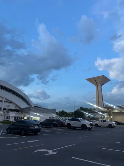 This media file shows an outdoor scene at dusk, with a clear blue sky filled with scattered clouds. The main subjects include a line of parked cars on a paved surface marked with white lines, suggesting a parking lot or street. The cars are of various colors, predominantly dark and white. Behind the cars, there's a modern building with a curved architectural design, featuring numerous windows and external air conditioning units. Some lights are visible on the building's facade, indicating artificial illumination.
Prominently featured in the upper portion of the image is a tall, distinctive concrete structure with a triangular, abstract shape, possibly a monument or a piece of public art. The background also reveals a line of trees and the continuation of the building complex. The overall setting appears to be urban or semi-urban.
The time of day is late evening, suggested by the dimming natural light and the activated artificial lights. The weather seems fair, with the sky being mostly clear blue despite the presence of clouds. There are no people visible in the frame.
A sign is partially visible on the building, with text in what appears to be Thai script. One visible line of text reads "มงคล" (Mongkol), which translates to "auspicious" or "lucky". This sign likely provides information related to the location, possibly indicating it is a place of significance or a public facility within Tha Khlong Town Municipality, Thailand. The image captures a tranquil and somewhat imposing atmosphere, highlighting the blend of modern architecture and a unique landmark against the twilight sky.
