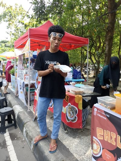 A young man with curly hair and white sunglasses pushed up on his forehead stands in an outdoor market, holding a white foam container of food and a small spoon. He wears a black t-shirt with "Menata" printed on it, light blue rolled-up jeans, and blue flip-flops, and his mouth is slightly open, as if speaking or about to take a bite.
The setting is a vibrant daytime street market, likely in Mataram, Indonesia, characterized by red canopy tents and various food stalls. Green trees provide shade in the background. On the left, another person in a pink hijab is partially visible near a stall. On the right, a person in a dark green hijab and niqab is seen bending over behind a stall. Black plastic stools are in the foreground, next to a concrete curb.
The stalls display various food items in clear containers, and banners are draped over tables. One red banner on the right stall features text: "0877-5503-1783," "@CHILLIFOOD_," and "CHILLIFOOD," with an image suggesting "Spaghetti." Another banner under the main subject displays an image of shellfish. In the far background, motorcycles are parked, common in such market settings. The scene conveys a bustling, casual atmosphere of street food vendors and patrons.