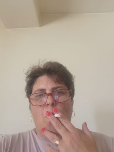 A woman with short, dark, tousled hair and red-rimmed glasses is holding a lit cigarette to her mouth. She is wearing a light-colored t-shirt and has bright red nail polish on her fingers. A silver hoop earring is visible on her left ear. The background is a plain, light-colored wall. The image appears to have been taken indoors, as there is a subtle shadow cast from a corner in the upper left portion of the frame, suggesting it is a room. The specific location within Lomets, Bulgaria, cannot be determined from the visual information provided.