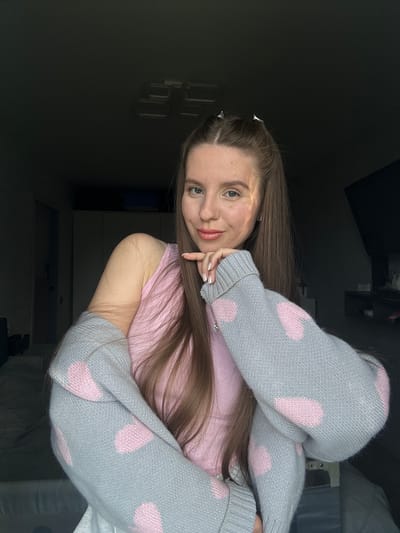 A young woman is reclining on what appears to be a bed or soft surface. She is wearing a light pink t-shirt and a light gray, long-sleeved sweater or pajamas with pink polka dots. Her long, straight brown hair is parted to the side. Her head rests on her hand, with her fingers near her mouth. The background includes a dark, shadowy area with reflections of what seems to be a television or window. The location, Klin, Russia, cannot be determined from the image alone.