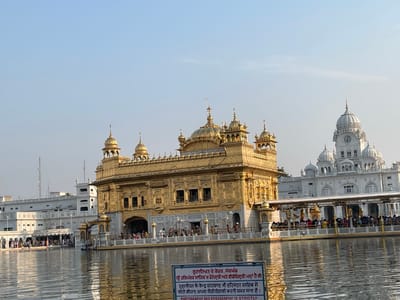 This image shows the Golden Temple, also known as Sri Harmandir Sahib, in Amritsar, India. The iconic golden structure, a prominent Sikh gurdwara, is situated in the center of a large, reflective body of water, known as the Amrit Sarovar or Pool of Nectar. The temple is adorned with intricate carvings and topped with golden domes and spires. To the right of the Golden Temple, a large white building with white domes and a clock tower is visible, part of the complex that surrounds the sacred pool.
In the foreground, a sign with text in Punjabi and English is placed in the water. The sign states that photography and videography are strictly prohibited. A wide walkway with a white railing extends from the shore to the Golden Temple, with a crowd of people walking along it. Many individuals are visible on the walkway, some standing and observing, while others are in motion. The water's surface is rippled, reflecting the light and the surrounding architecture. The sky above is clear and light blue, indicating daytime. The overall scene depicts a serene and spiritual atmosphere at this important religious site.