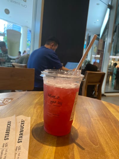 A cold, pink beverage in a clear plastic cup with a straw sits on a wooden table in a Starbucks cafe. The cup has a handwritten message that reads "Believe in yourself". Two Starbucks paper sleeves are laid out on the table in front of the cup. In the background, several people are seated at tables and working on laptops. The cafe appears to be well-lit with natural light coming in from a large window. The location is identified as Starbucks, West Jakarta, Indonesia.