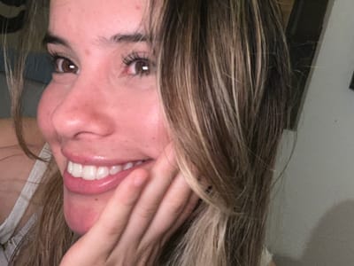 A close-up, upside-down selfie of a fair-skinned person with brown eyes and light brown hair with blonde highlights. The person's right hand is covering part of their face, near the mouth. They are smiling, revealing white teeth and pink lips. A small portion of a white tank top is visible on the right side of the frame. The background is blurry, suggesting an indoor setting, with glimpses of a white wall and a dark object. It is impossible to determine the location of Sumalao, Argentina from this image alone.