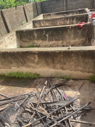 In Kakau, Nigeria, a series of tiered concrete basins are visible, possibly remnants of a former industrial or agricultural facility. The basins are weathered and show signs of disrepair, with some vegetation growing along their edges. In the foreground, a pile of dry, burnt vegetation and debris lies on the concrete floor, suggesting recent or past fires. A red cloth is draped over the edge of one of the basins in the middle ground, and various containers, including a red plastic basin and metal bowls, are placed on the edge of the furthest basin. The background features a brick wall and lush green foliage, indicating an outdoor setting with some overgrowth. The scene appears to be an abandoned or neglected area within the city.