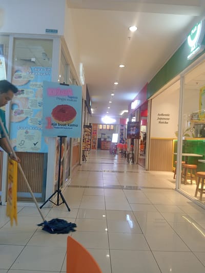 This media file captures a view down a brightly lit shopping mall corridor. The foreground shows a person in a blue shirt mopping the tiled floor with a blue mop. To the left, a yellow caution sign is visible.
Dominating the left side of the image is a large poster with the text "Apa Sih Bedanya??" (What's the difference??) and several images of donuts, along with descriptions in Indonesian. Next to it, another poster features the word "Diet" in large pink letters, followed by "Tapiiiiiii Pingin makan" (But still want to eat) and an image of a single donut. Below the donut, "aja buat kamu" (just for you) and "#happy diet mulai besok" (#happy diet starts tomorrow) are displayed.
Further down the corridor, shops line both sides. One shop on the right has a sign that reads "Authentic Japanese Matcha". Several other shop signs and displays with text in Indonesian are visible, including what appears to be a food stall with prices like "12K" and "6K". Red and orange chairs are set up outside some of the establishments.
The overall scene suggests a commercial environment, likely during operating hours, given the lighting and activity. The focus on donuts and diet in the posters introduces a lighthearted or humorous element. The clean, tiled floor indicates recent maintenance. The setting is an indoor public space, characteristic of a shopping mall in Surabaya, Indonesia.