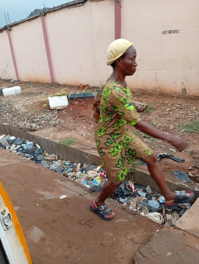 An older woman, wearing a yellow hat and a green and yellow patterned dress, is walking across a narrow, concrete-lined drainage ditch filled with trash in Ogidi, Nigeria. She is wearing sandals. The ditch is located next to a dirt path, and a painted wall runs in the background. The writing "WEST N'ODILI" is visible on the wall.