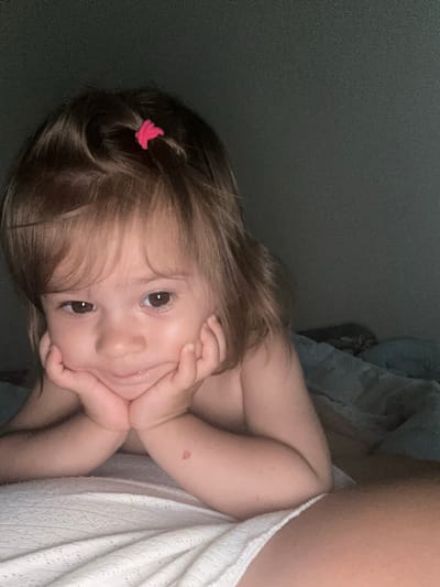 The media file shows a close-up, vertically oriented, indoor shot of a young child lying down, possibly in bed. The child has light brown hair, with a small pink hair tie on the side. Their face is turned towards the camera, with hands resting near their chin, suggesting a relaxed or contemplative pose. The child's eyes are open and looking directly at the viewer. A faint redness, possibly a birthmark or a small bruise, is visible on their arm. The background is dark and out of focus, with a hint of a light-colored textured fabric, perhaps a blanket, to the right. The lighting appears to be dim, suggesting it could be nighttime or a dimly lit room. No text is visible in the image.