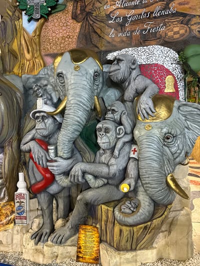 The image depicts a large, elaborate sculpture in Alicante, Spain, featuring a group of gray, realistic-looking chimpanzees and two elephants. The sculpture is part of a larger mural or installation that includes painted elements and text.
In the foreground, several chimpanzees are arranged in a cluster. One chimpanzee, wearing a white hat and holding a red, curved object and a metallic cup, sits prominently. Another chimpanzee is positioned above and behind him, and a third is nestled on the elephant's trunk. A smaller infant chimpanzee is depicted curled up on the elephant's tail. A red cross emblem is visible on one of the chimpanzee's arms.
Two elephants are integrated into the composition. The head of one elephant is to the right, with a golden headdress. Another elephant's trunk and part of its head are on the left. Both elephants have golden accents on their heads and tusks.
Scattered around the base of the sculpture are several objects. A white bottle with a label reading "ANIS TENIS" and "DESDE 1888" is on the left. To the right, a yellow sign with text in Spanish, titled "Guardianes del Tiempo ENFERMERIA," is visible. Another yellow, folded document with similar text is positioned to the left of the "ANIS TENIS" bottle.
The background of the image includes painted imagery and text. White and red mosaic tiles are present behind some of the figures. A portion of a painted mural depicts figures and various patterns. Text in white cursive script reads, "en Alicante, la Los Gorilas llenaba la vida de Fiesta." (In Alicante, the Gorillas filled life with Fiesta.)
The overall environment appears to be an indoor setting, possibly a museum or a public display area, with a floor covered in small white pebbles. The lighting suggests an artificial light source. The sculpture's style is detailed and somewhat whimsical, blending realistic animal forms with fantastical elements.