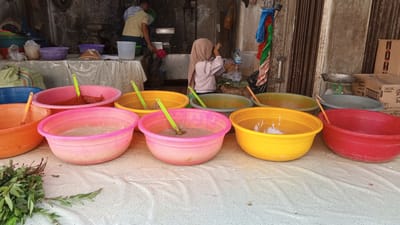 An outdoor market stall in Puloara, Indonesia, displays an array of colorful plastic bowls filled with various food preparations. In the foreground, a bunch of green leafy herbs lies on a white patterned tablecloth. Behind the herbs, a row of large, brightly colored bowls – orange, pink, yellow, and red – are arranged. Some bowls contain what appears to be a reddish paste, while others hold a lighter-colored, soupy mixture. Green plastic spoons are visible in several of the bowls.
In the background, a young girl wearing a light pink hijab and a light purple shirt stands at a table. She is facing away from the camera and appears to be handling something on the table, possibly preparing food. To the left of the girl, an adult, presumably a man, is also present at the stall, wearing a yellow shirt and a checkered head covering. He is facing the girl and holding a red container. Behind them, shelves are visible with various containers and items, suggesting a food preparation or vending area. The overall environment appears to be an open-air market with a rustic, possibly weathered, background structure. Boxes are stacked to the right, one labeled with "Royco FOODS."