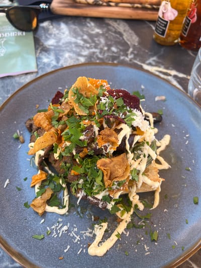 A close-up, overhead shot captures a dish of food served on a dark gray plate. The plate rests on a dark, marble-patterned table. The food is piled high, appearing to be a mix of sliced meat, root vegetables like beets, and crispy potato or vegetable chips, all garnished with chopped fresh green herbs and grated white cheese. A creamy white sauce is drizzled over the entire dish. In the background, a pair of dark sunglasses rests on a green booklet, and two small bottles of condiments are visible. The overall scene suggests an outdoor dining setting, possibly in the city of Tønsberg, Norway, given the context. The lighting is natural and the focus is sharp on the food.