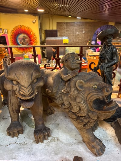 This is a medium-shot, eye-level, interior photograph of a Mexican craft shop or restaurant in Cuernavaca, Morelos, Mexico. The foreground is dominated by two carved wooden sculptures of mythological creatures, possibly a jaguar and a lion. The jaguar-like creature on the left is standing on four paws, with its mouth open and fangs exposed. The lion-like creature on the right is in a similar pose, with its mouth open and a smaller, human-like figure riding on its back. The sculptures are made of dark wood and show signs of age and wear. Behind the sculptures, a red metal railing separates the foreground from the background. The background depicts a colorful mural on a yellow wall, featuring a sun-like design. Further in the background, a statue of a man in a sombrero and traditional Mexican attire stands to the right. The environment appears to be a lively establishment, possibly a restaurant, with tables and chairs visible in the blurred background. The ceiling is constructed of wooden slats.