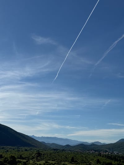 A high-angle, wide shot captures a sprawling, verdant landscape under a clear blue sky. Two prominent contrails streak diagonally across the sky from the top right towards the center left, with a fainter contrail visible lower down. The foreground is dominated by rolling green hills covered in dense vegetation, with a dark, steep hill occupying the left side of the frame. In the midground, a small, clustered village with red-tiled roofs is nestled amongst the trees. The background features layers of hazy blue mountains receding into the distance. The scene is devoid of any human presence or discernible activity.