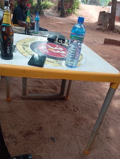 The scene depicts an outdoor gathering in Okpatu-Ukehe, Nigeria. A rectangular table with a white top and yellow plastic trim, supported by grey metal legs with yellow plastic caps, occupies the foreground. On the table, from left to right, are a brown glass bottle of Guinness Foreign Extra Stout, a black smartphone lying screen-down, a circular coaster displaying a compass rose and "STAR" branding, two black flip phones, small metallic items (possibly bottle openers or keys), and a clear plastic bottle of Mt Salva table water with a blue cap. In the midground, two men are partially visible seated behind the table. Both men wear dark shirts and red caps. Another green beer bottle and a clear water bottle with a blue cap are also visible on the table near them. The ground is reddish-brown sandy earth with visible footprints and minimal debris. The background features sparse green vegetation, an upward sloping terrain, and a visible reddish-brown structure and tree trunk on the right. The scene is illuminated by natural daylight.