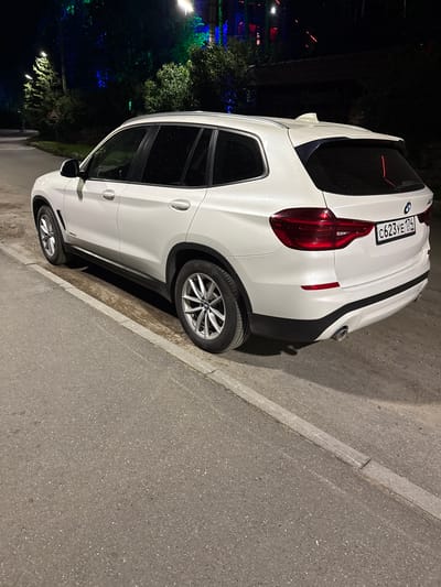 A white BMW SUV is parked on the side of a paved road at night in Turgoyak, Russia. The car, a BMW X3 model, is facing away from the viewer and is positioned diagonally across the frame, with its rear end closer to the camera. The vehicle has tinted windows and a license plate reading "C623YE 174 RUS". The road appears to be in an urban or semi-urban area, with a concrete curb bordering the asphalt. In the background, trees and lights are visible, suggesting the presence of buildings or a park. The overall lighting is dim, typical of nighttime conditions, with some artificial lights casting reflections on the wet surface of the car, indicating recent rain. There are no people clearly visible in the immediate vicinity of the car, though distant figures are present further down the road. The scene is static and appears to be an ordinary street view at night.