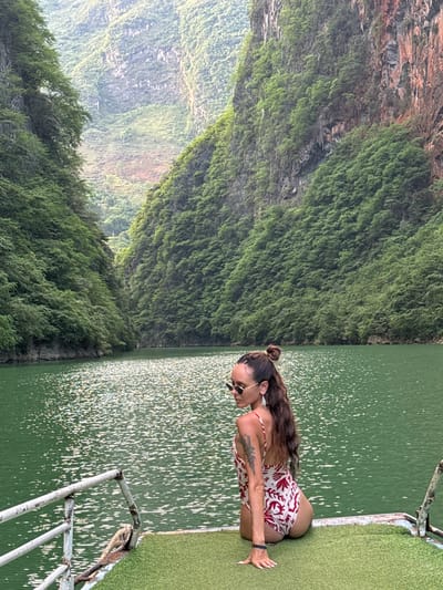 An eye-level shot captures a woman in a patterned swimsuit, kneeling at the edge of a platform adjacent to a body of water. The woman has long brown hair styled in a bun, and she wears sunglasses. A tattoo is visible on her upper arm. The platform has artificial green turf. The water surface glitters with reflected light. Across the water, a steep, verdant hillside dominates the frame. The location is Đồng Văn Commune, Vietnam.