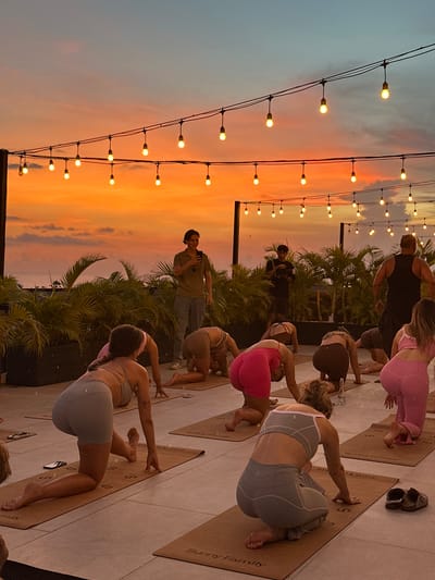 The image depicts a group of people participating in an outdoor yoga session at dusk, likely in Tibubeneng, Indonesia. The sky exhibits a vibrant orange and pink sunset. A string of lights hangs above the scene, illuminating the yoga participants. Most people are actively engaged in yoga poses, while a man in a green shirt stands amongst them, possibly instructing or observing. The yogis are positioned on individual yoga mats. Lush tropical vegetation, including palm trees, is visible in the background. Some discarded footwear is evident on the surface where the yoga mats are placed. The overall setting suggests a rooftop or elevated platform overlooking the landscape.