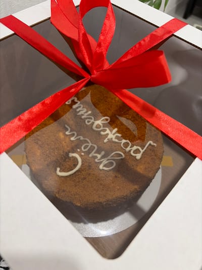 A cake inside a white square box is tied with a red ribbon. The cake appears to be brown with white frosting in a script. The box is sitting on a surface that looks like a floor or table. The frosting reads "С днём рождения" which translates to "Happy Birthday." The box is a see-through box. The exact location in Sarapul, Russia cannot be determined from the image.