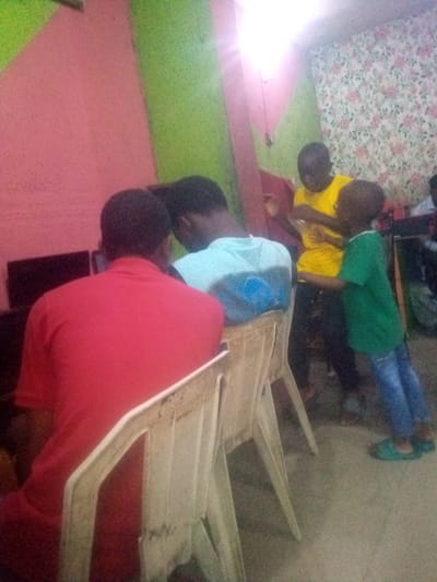 In a room in Shangisha, Nigeria, three young males and one adult male are engaged in activities. One young male, wearing a yellow t-shirt, stands and gestures. Another young male, in a green t-shirt and blue jeans, stands facing him. In the foreground, an adult male in a red shirt is seated at a computer, with his back to the camera. To his left, another male in a light blue t-shirt is also seated, facing the computer. The room has pink and green walls and floral wallpaper.