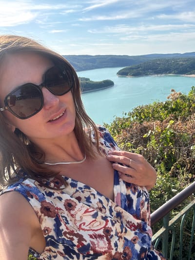 In Moirans-en-Montagne, France, a woman wearing large, dark sunglasses and a floral dress poses for a selfie against a backdrop of a bright blue lake surrounded by lush, green hills under a partly cloudy sky. She has brown hair and is wearing a silver necklace. Her right hand is near her chest, and she has a ring on her right ring finger. The woman is standing next to a railing with green metalwork, suggesting she is on a balcony or viewpoint. The scene is set outdoors, likely on a sunny day.